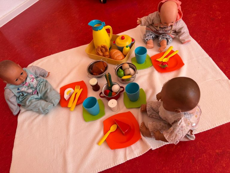 Essen und Trinken Drei Puppen sitzen um einen Tisch mit buntem Geschirr und Spielessen. Mittagessen, Fesper, Gesunde Ernährung